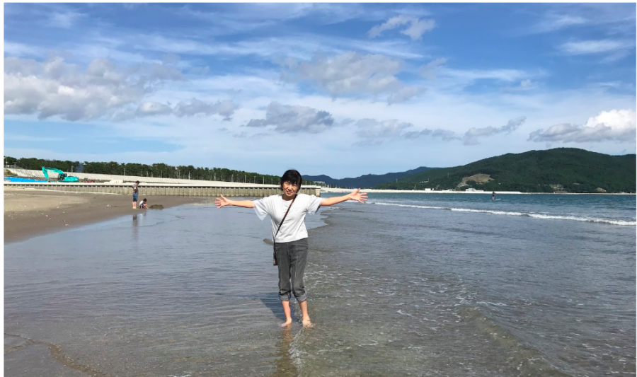The writers grandmother posing on the beach in Ishinomaki. On the far left there is a green excavator and the white horizontal line along the center of the picture is the dike.